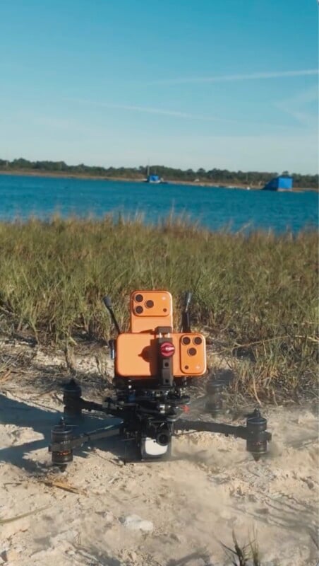A drone equipped with several orange smartphones is positioned on sandy ground near tall grass, with a body of water and blurred blue structures in the background under a clear sky.