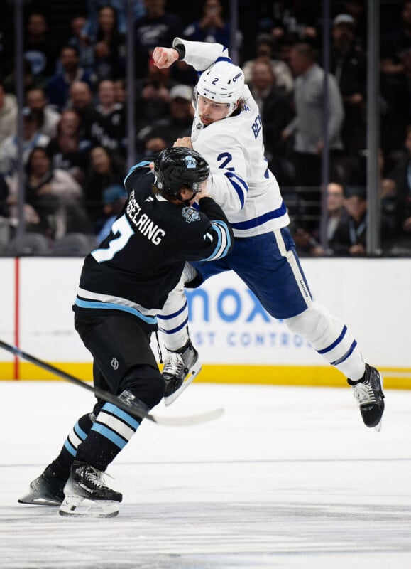 A hockey player in a white uniform jumps and collides mid-air with a player in a black uniform during a game, while spectators watch from the stands in the background.