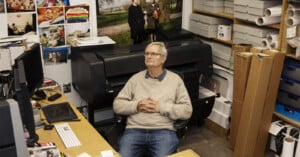 An older man in glasses and a beige sweater sits in an office surrounded by computers, printers, photos on the wall, and shelves filled with boxes and paper rolls. He looks slightly upward with his hands clasped.