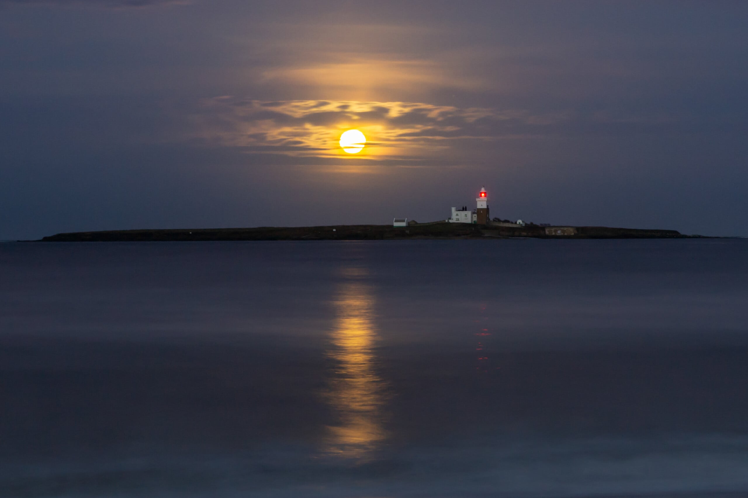 Full moon rises behind a small island with a lighthouse, casting a yellow reflection across calm water under a dark, cloudy sky. The lighthouse’s red light is visible in the distance.