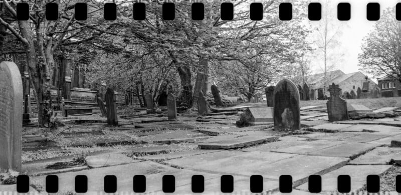 Black-and-white photo of an old graveyard with weathered headstones and stone pathways, surrounded by leafless trees. A building is visible in the background. Film sprocket holes frame the image.
