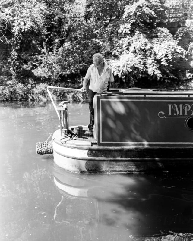 A man stands on the back deck of a canal boat on a calm river, surrounded by leafy trees. Sunlight filters through the branches, casting dappled shadows on the water and the boat.