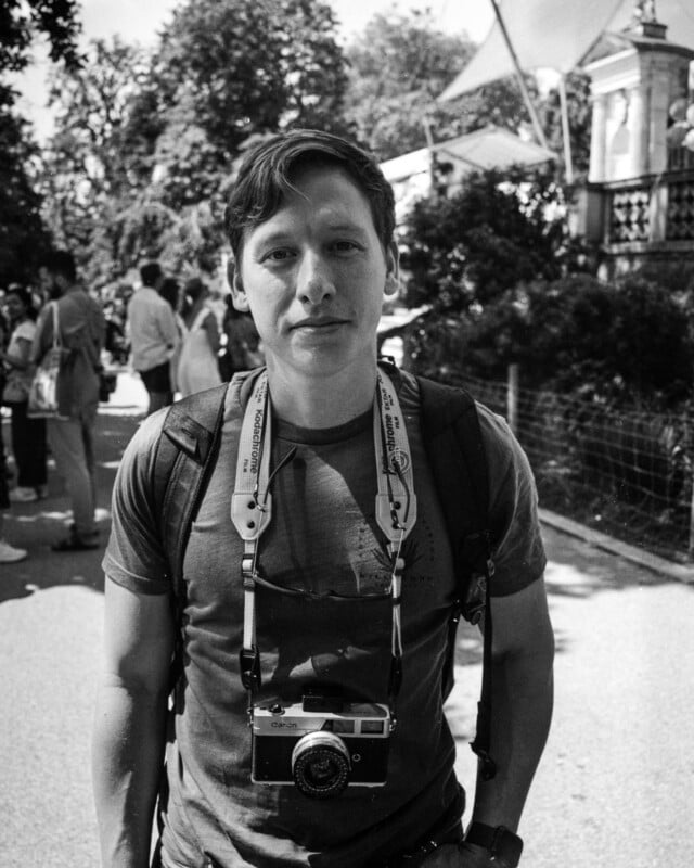 A man stands outdoors wearing a backpack and a camera around his neck. He looks at the camera, with trees, people, and part of a building in the background on a sunny day. The image is in black and white.