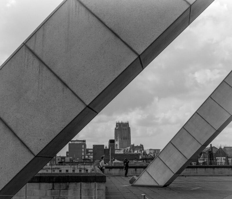 Two large, diagonal concrete beams frame a cityscape with a cathedral in the background. Two people are seated on a low wall in the middle ground, and the sky is cloudy. The image is black and white.