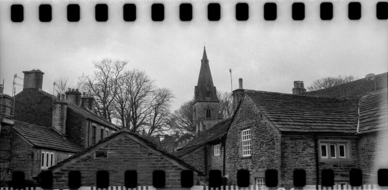 Black-and-white photo of stone buildings with sloped roofs, bare trees, and a tall church steeple in the background. The image is framed by the borders of a strip of 35mm film.