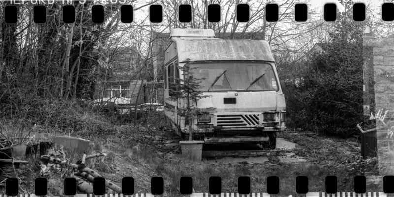 A black and white photo of an old, weathered camper van parked in an overgrown yard, surrounded by trees and bushes. The image includes film sprocket holes along the edges.