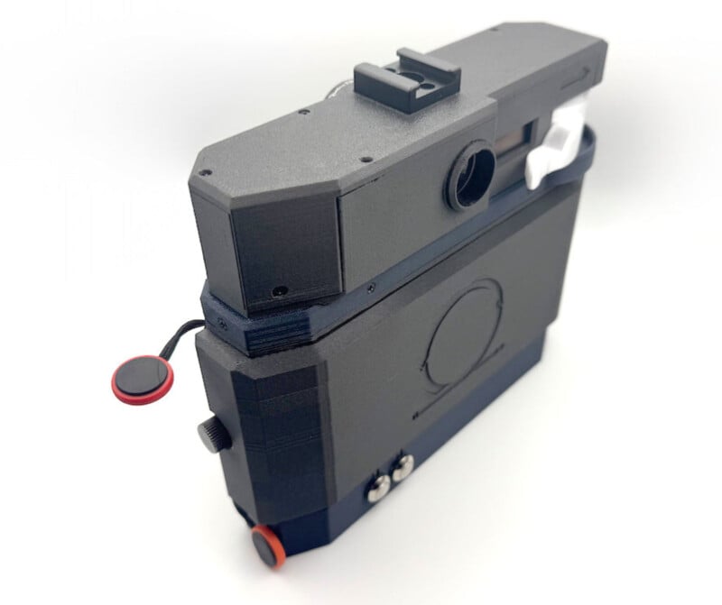 A black, boxy, retro-styled camera with angular edges, a round lens opening, red and black buttons, and a prominent viewfinder, photographed on a white background.