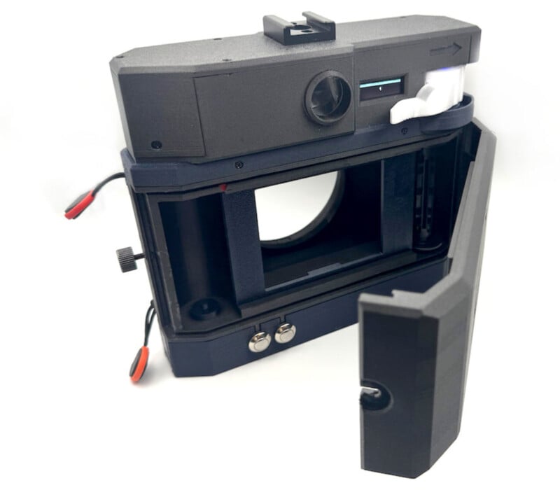A close-up of a black and dark blue film camera with its back cover open, revealing the film compartment and internal components. Wires and buttons are visible on the side and bottom.