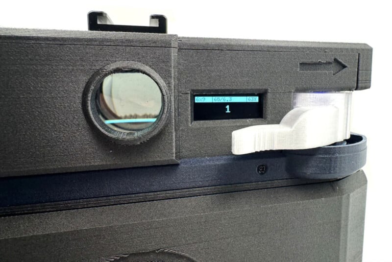 Close-up of a black 3D-printed camera with a round lens, a small digital display showing "1", and a white control dial on the right. An arrow is embossed above the dial on the camera's top right.