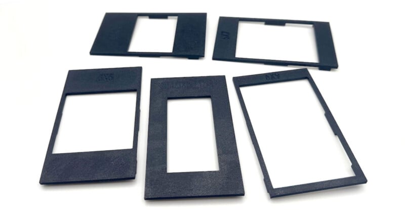 Five black rectangular film slide mounts with large cut-out windows are arranged on a white surface. Three mounts are in the front row and two are in the back row.