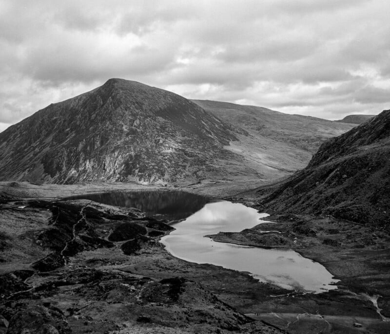 Black and white photo of a rugged mountain landscape with a large hill, a reflective lake in the foreground, rocky terrain, and cloudy skies above.