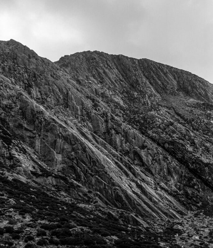 Black and white photo of a rugged, steep mountain slope with rocky textures and sparse vegetation under a cloudy sky.