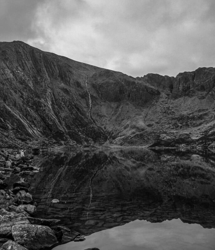 Black and white photo of a rocky mountain reflected in a still lake, with cloudy skies above. The shoreline is covered in rocks, and the scene feels calm and remote.