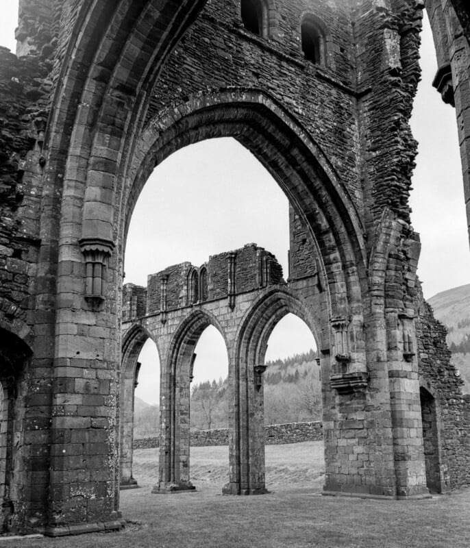 Black and white photo of large stone arches and ruins of an old abbey or church, showing weathered, detailed masonry, with grassy grounds and hills visible through the arches in the background.