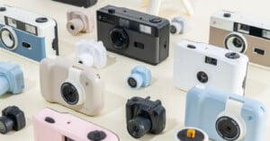 A variety of retro-style cameras in pastel shades of blue, beige, white, and black are displayed on a light surface, with both regular-sized and mini cameras arranged closely together.