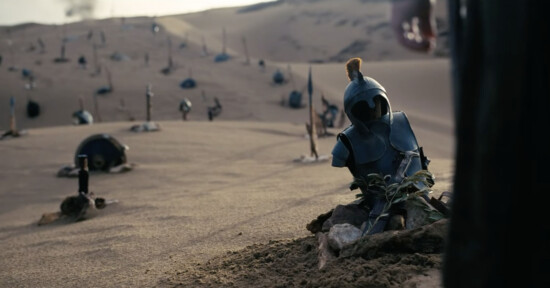 A desert landscape with scattered helmets and armor pieces on sand mounds, resembling makeshift graves. In the foreground, a blue helmet and chest plate are placed among stones and small branches.