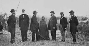 A historical black-and-white photo shows six men and one woman standing outdoors in a field, dressed in 19th-century attire. The men wear coats and hats, and the woman is wrapped in a dark cloak.