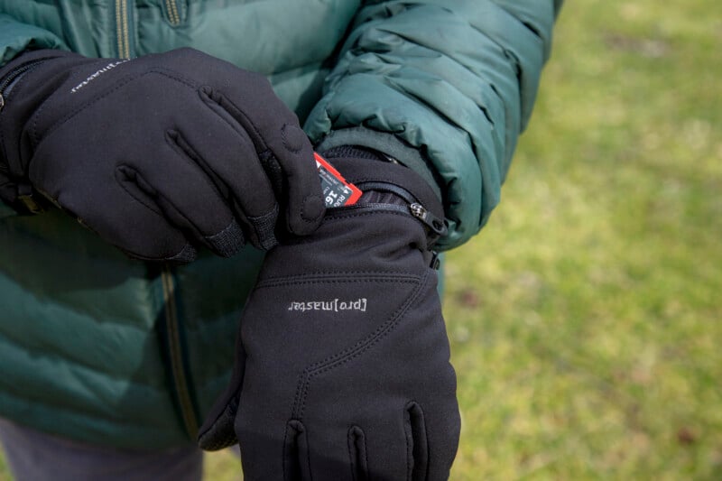 A person wearing black gloves and a green jacket inserts a red card into a small zippered pocket on the back of one glove while standing on grass.