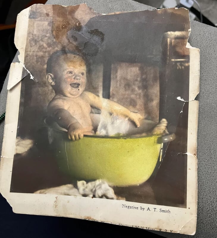 A vintage, worn photograph of a smiling baby sitting in a yellow wash basin filled with soapy water, with visible creases and damage on the photo. The text “Negative by A. T. Smith” appears at the bottom.