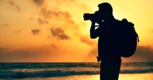 Silhouette of a person with a backpack taking a photo with a camera on a beach at sunset, with orange and yellow hues in the sky and the ocean in the background.
