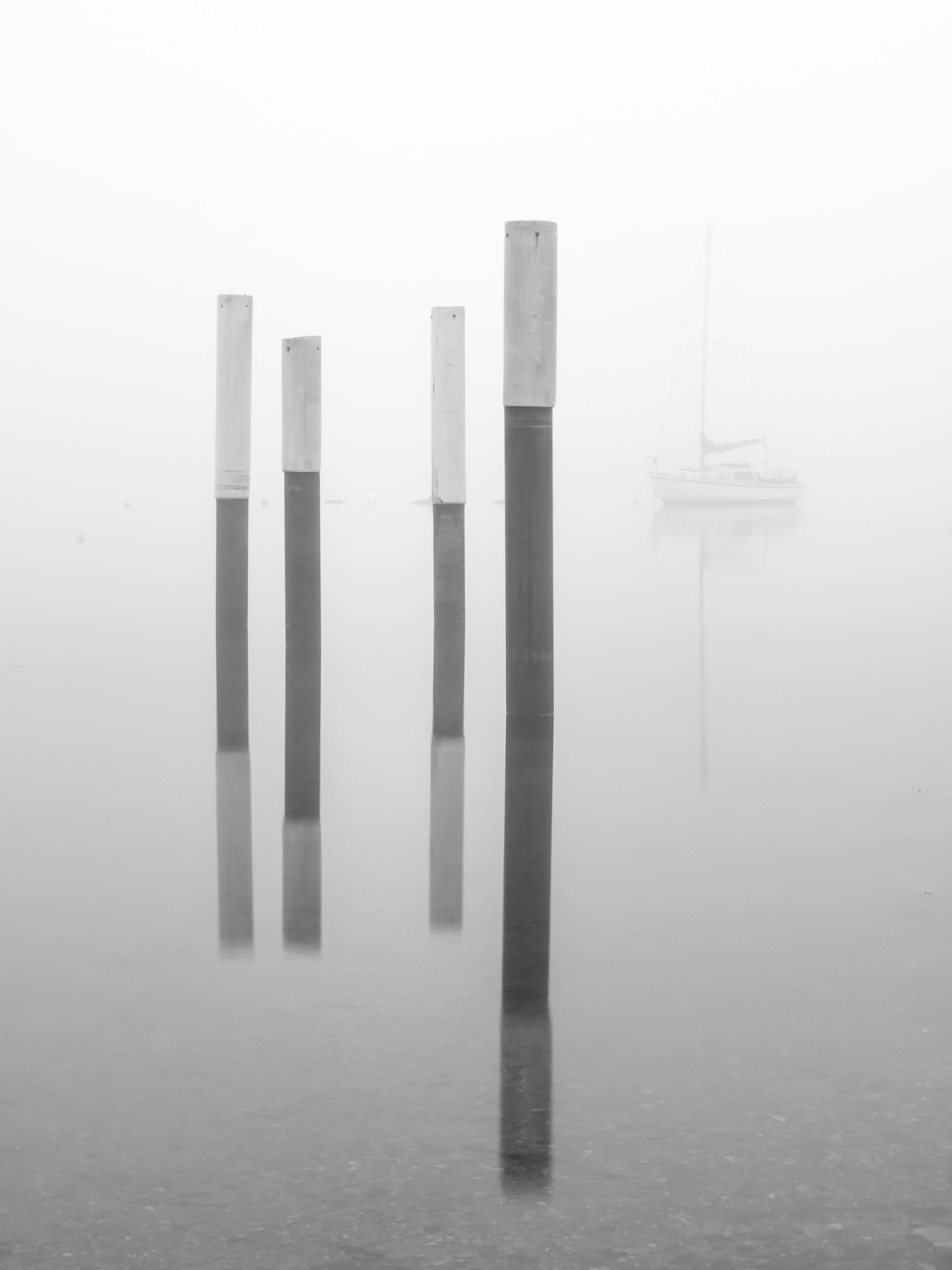 Cuatro pilares verticales del muelle se alzan en el agua tranquila, sus reflejos visibles. A través de la espesa niebla, un velero se ve apenas al fondo, creando una escena tranquila y minimalista.