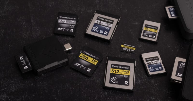 Varias tarjetas de memoria ProGrade de varios tamaños y capacidades están dispuestas sobre una superficie negra, junto a un lector de tarjetas USB negro. Las tarjetas muestran varias etiquetas de velocidad y almacenamiento en amarillo y gris.