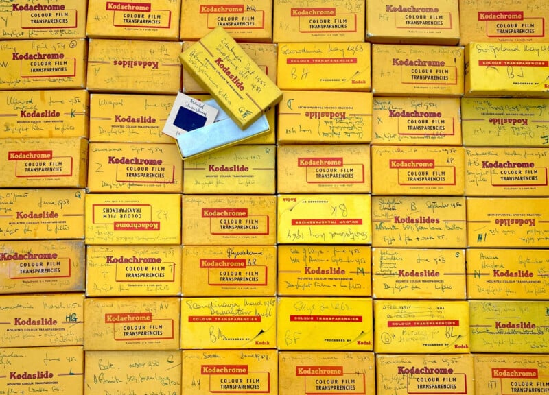 Pilas de cajas de diapositivas Kodachrome amarillas, muchas de ellas con etiquetas escritas a mano. Se abrió una caja con una diapositiva azul en su interior. Los cuadros llenan toda la imagen, creando un colorido patrón vintage.