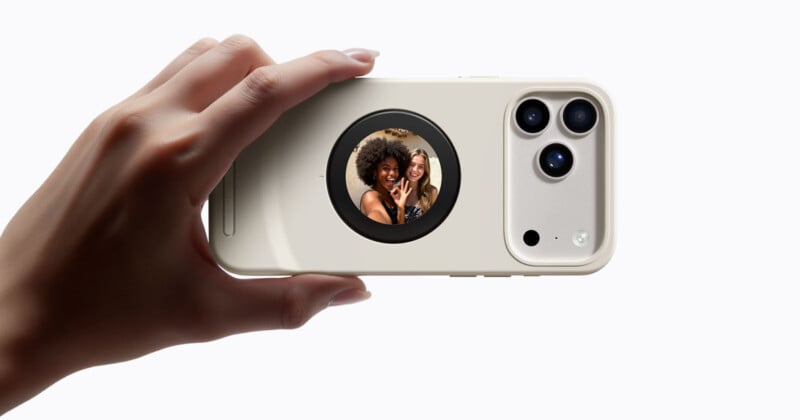 En una mano sostiene un teléfono inteligente con carcasa beige y una pequeña pantalla circular en la parte posterior, que muestra a dos mujeres sonriendo y haciendo el signo de la paz para una selfie.