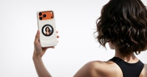 A woman with short, wavy hair and a black sleeveless top holds up a smartphone with a circular display on the back showing her own image, as if taking a selfie.