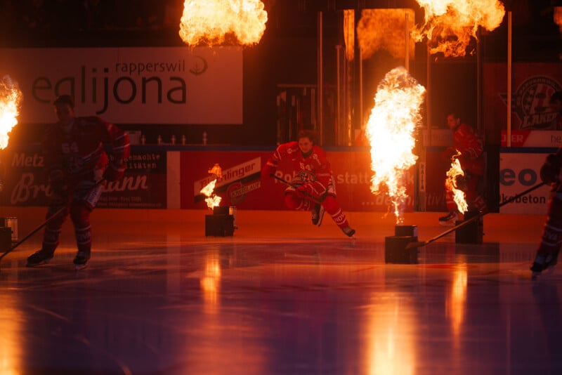 En la espectacular entrada a la arena, los jugadores de hockey se deslizan sobre el hielo a través de columnas de fuego ardiente, con un jugador saltando y toda la escena iluminada por llamas.