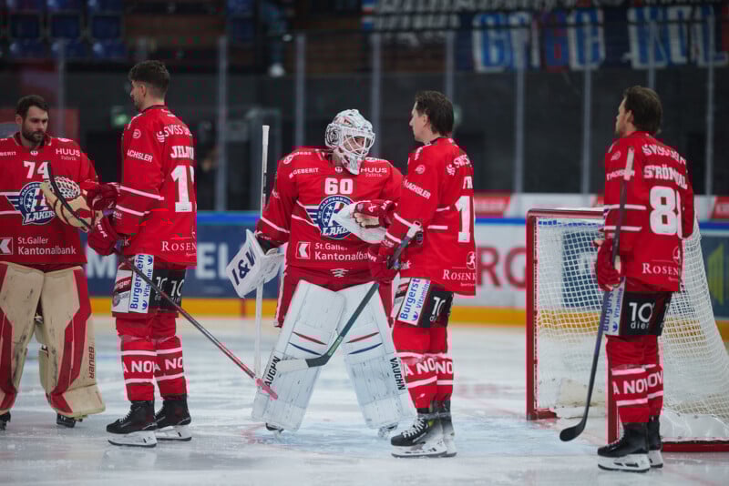 Cuatro jugadores de hockey con uniformes rojos y un portero con todo su equipo están de pie sobre el hielo, charlando y sosteniendo palos de hockey. Al fondo se muestra un estadio con asientos azules vacíos y publicidad.