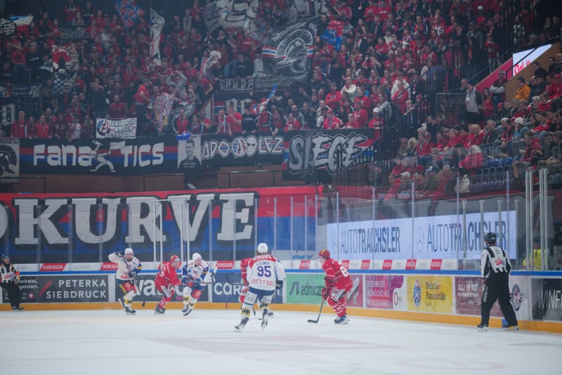 Los jugadores de hockey sobre hielo juegan en la pista de hielo con un árbitro cerca. Las gradas estaban repletas de aficionados vestidos con camisetas rojas ondeando banderas y pancartas, y el ambiente era animado y animado.