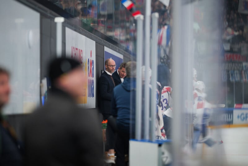 Dos hombres trajeados se encuentran cerca del cristal de una pista de hockey sobre hielo, con jugadores y espectadores claramente visibles en primer plano y al fondo. Un hombre es calvo y ambos parecen estar observando el juego.