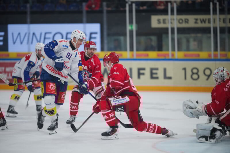 Los jugadores de hockey con uniformes rojos y blancos compiten por el disco frente a la portería, con el portero vestido de blanco preparándose para bloquear; Al fondo se puede ver una arena con espectadores.