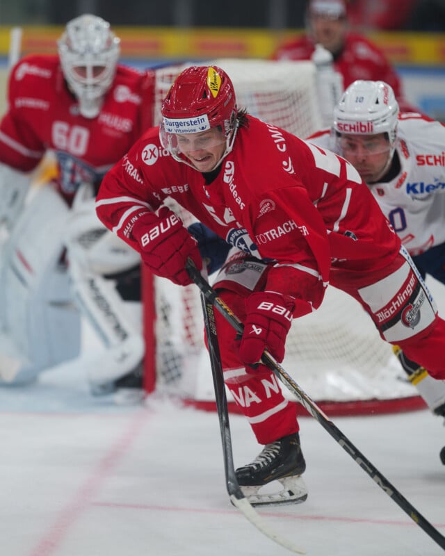 Un jugador de hockey con uniforme rojo patina hacia adelante con el disco completamente enfocado, mientras su oponente con uniforme blanco lo persigue. El portero vestido de rojo está alerta al fondo, cerca de la portería.