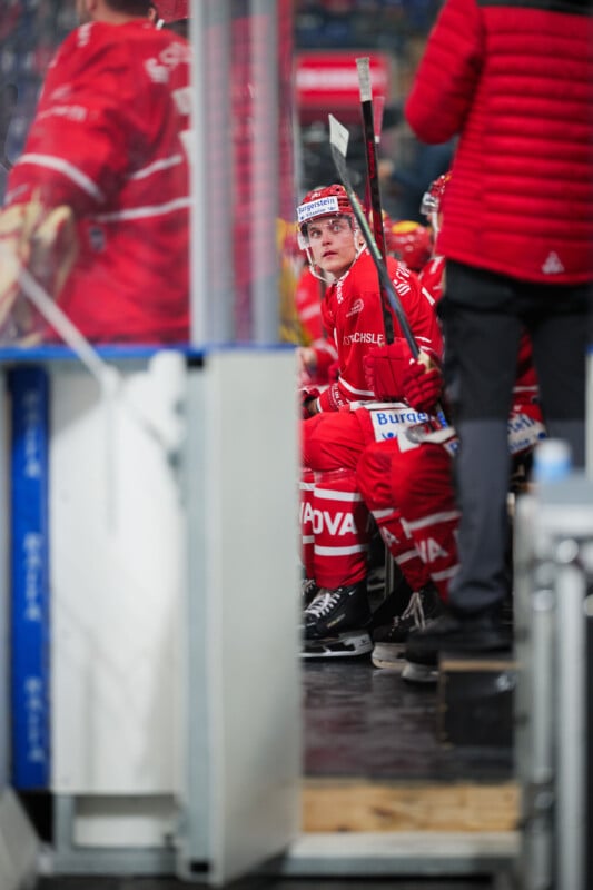 Un jugador de hockey con uniforme rojo y casco está sentado en un banco, sosteniendo un palo y mirando hacia arriba, mientras otros jugadores con uniformes rojos están cerca. La escena se desarrolla dentro de una pista de hockey sobre hielo.