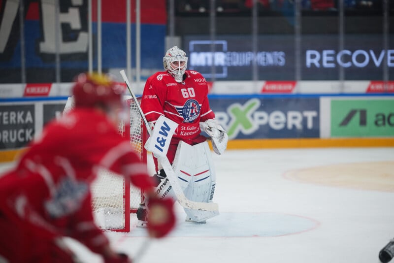 Un portero de hockey sobre hielo vestido con ropa roja y blanca está alerta en la red mientras otro jugador patina en primer plano, ambos con uniformes rojos. En los carteles de la pista se muestran varios anuncios.
