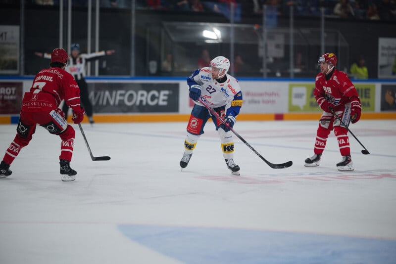 Tres jugadores de hockey sobre hielo jugando en la pista de hielo. Un jugador con camiseta blanca con detalles en azul y amarillo maneja el disco, mientras dos jugadores con camisetas rojas se acercan a él. Al fondo se ve un tablero de ajedrez y espectadores parcialmente visibles.