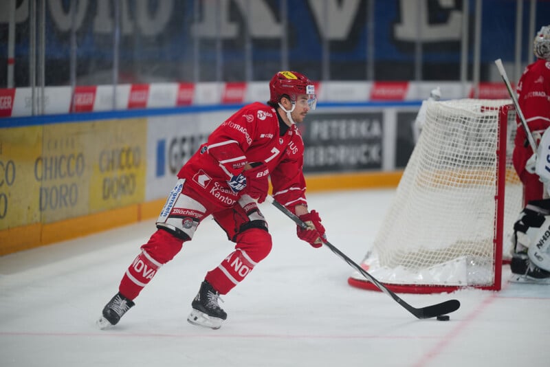 Un jugador de hockey con uniforme rojo patina sobre el hielo cerca de la portería, controlando el disco con su palo, con el portero y la red visibles al fondo.