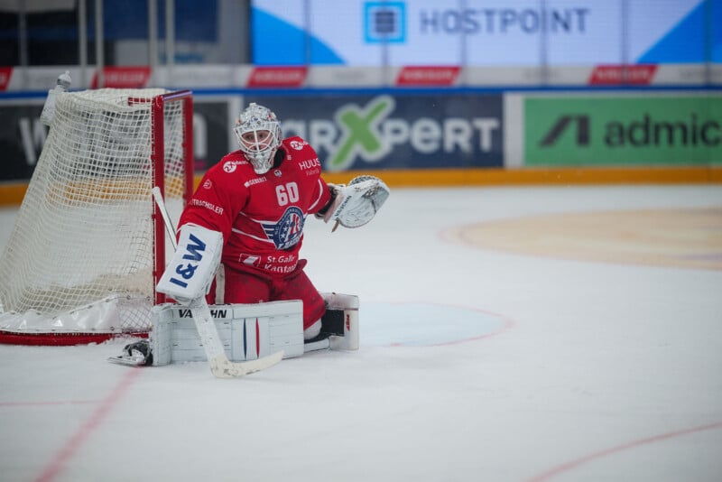Un portero de hockey vestido con ropa roja y blanca se arrodilla sobre el hielo frente a la portería, con el guante en alto, observando el partido. Al fondo se muestran anuncios a lo largo de la pista de hielo.