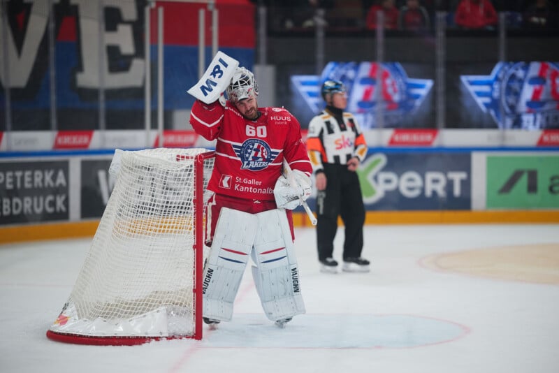 Un portero de hockey vestido con ropa roja y blanca se encuentra junto a la red, sosteniendo su máscara. Al fondo, sobre el hielo, hay un funcionario vestido de blanco y negro. La publicidad del estadio y la multitud borrosa detrás son claramente visibles.
