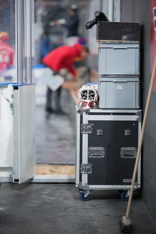 Una máscara de portero de hockey se encuentra encima de una gran caja de engranajes negra con ruedas con dos cajas grises apiladas encima. Al fondo, un hombre con una chaqueta roja se difumina cerca de un banco de pista. Una escoba está apoyada contra la pared.