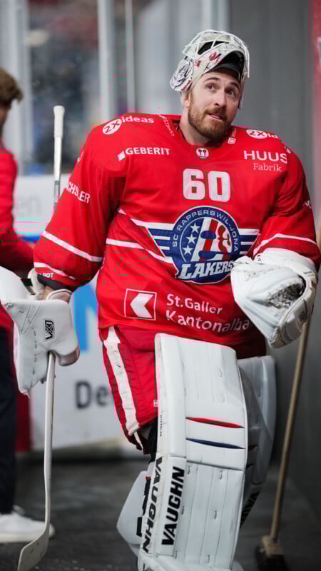 Un portero de hockey con una camiseta roja de los Lakers y el número 60 se encuentra en una pista cubierta sosteniendo un palo y un casco, usando protecciones blancas de portero y con barba.