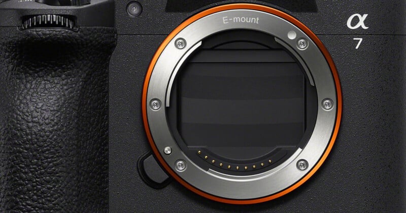 Primer plano del cuerpo de la cámara Sony Alpha 7, que muestra el conector de la lente con montura E, el anillo metálico naranja y los contactos eléctricos (sin la lente colocada). El mango de la cámara y la textura son visibles a la izquierda.