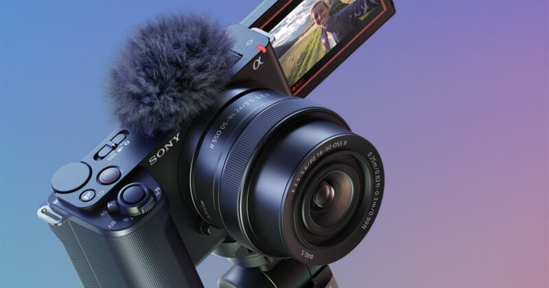 Cámara digital Sony con accesorio de micrófono mullido montado sobre trípode. Una pantalla plegable muestra a una persona tomándose una selfie al aire libre, con campos verdes de fondo.