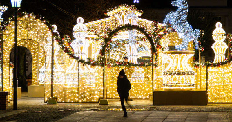 Un hombre vestido con ropa de invierno pasa junto a decoraciones festivas de luces amarillas y blancas brillantes, incluidos arcos y fuentes, en una calle de la ciudad por la noche. Un hombre la mira desde atrás.