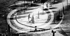 A black-and-white photo showing two blurry figures walking or cycling on a curved path, overlaid with a semi-transparent white swirl graphic. Sparse trees and poles line the empty, sunlit area.