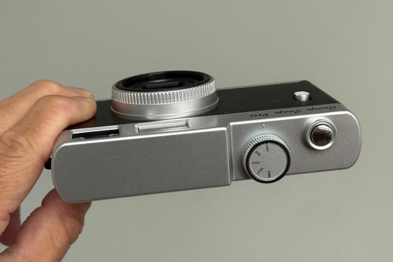 A hand holds a compact, silver digital camera viewed from the top, showing a lens, control dial, and button. The words "Camp Snap Pro" are inscribed near the edge. The background is plain and light gray.