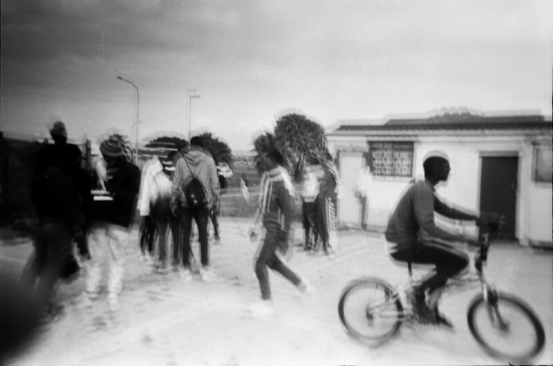 Una fotografía en blanco y negro muestra a un grupo de personas caminando juntas, con una persona en bicicleta en primer plano. La imagen está borrosa, dando sensación de movimiento. Al fondo se ven claramente los edificios y las farolas.