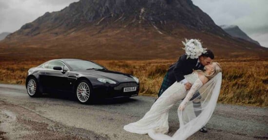 A groom dips and kisses his bride, who holds a bouquet and wears a long veil, on a road in front of a black sports car and a large mountain in a scenic, grassy landscape.